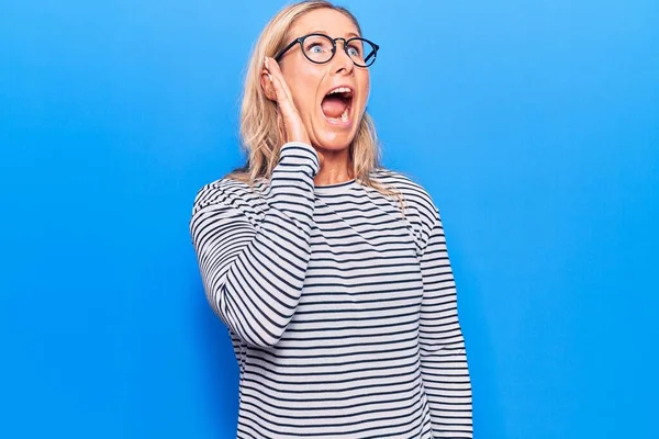 Middle age caucasian blonde woman wearing casual striped sweater and glasses shouting and screaming loud to side with hand on mouth. communication concept.