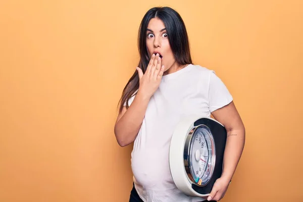 Young beautiful brunette pregnant woman holding controlling weight using weighting machine covering mouth with hand, shocked and afraid for mistake. Surprised expression