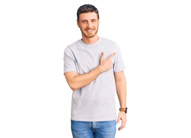 Yüzünde mutlu ve doğal bir ifadeyle, elleri ve parmaklarıyla yüzünü gösteren, gülen, neşeli tişörtlü, ayı gibi yakışıklı genç bir adam. 