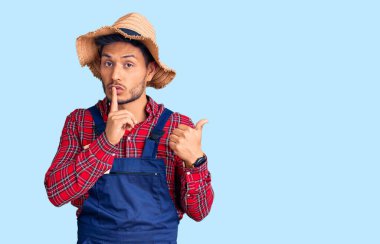 Yakışıklı Latin Amerikalı, tamirci üniforması giyen, sessiz olmak isteyen, parmakları dudaklarında, elleri yandan gösteren genç bir adam. sessizlik ve gizli konsept. 