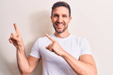 İzole edilmiş beyaz arka plan üzerinde duran sıradan tişört giyen genç yakışıklı adam gülümsüyor ve kameraya bakıyor iki eliyle ve parmaklarıyla kenarı işaret ediyor..