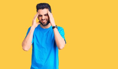 Sakallı, elleri kafasında, günlük tişört giyen genç yakışıklı bir adam. Stres yüzünden baş ağrısı çekiyor. migren ağrısı. 