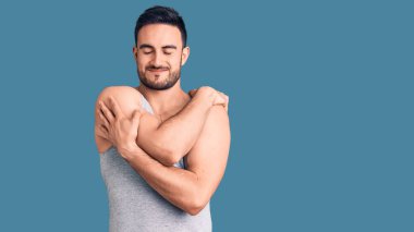 Mayolu ve kolsuz tişörtlü genç yakışıklı bir adam mutlu ve pozitif, kendinden emin bir şekilde kendine sarılıyor. Sevgi ve özgüven. 