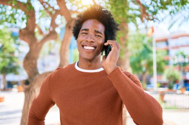 Genç, yakışıklı, Afro-Amerikan bir adam günlük giysiler giyip mutlu gülümsüyor. Yüzünde tebessümle dikilip şehrin caddesinde akıllı telefondan konuşuyor..