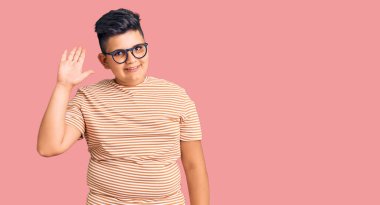 Günlük kıyafetler ve gözlük takan küçük çocuk merhaba diyor mutlu ve gülümsüyor, dostça karşılama hareketi yapıyor. 