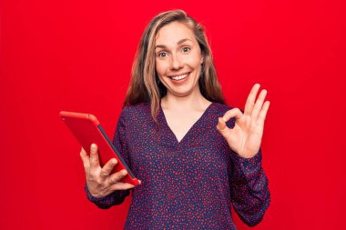 Güzel sarışın kadın, izole edilmiş kırmızı arka plan üzerinde tablet kullanıyor. Parmaklarıyla işaret veriyor, gülümsüyor. Mükemmel bir sembol. 
