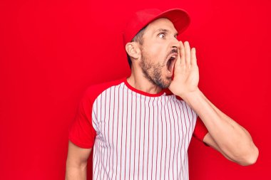Genç yakışıklı oyuncu beysbol kıyafetleri giyip izole edilmiş kırmızı arka planda el ele bağırıyor ve ağızdan ağıza bağırıyordu. İletişim konsepti.