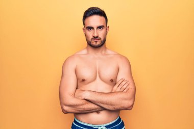 Sakallı, kolsuz tişörtlü genç yakışıklı adam sarı arka plan üzerinde duruyor şüpheci ve gergin, yüzünde çapraz kollu tasvip etmeyen bir ifade. Negatif kişi.
