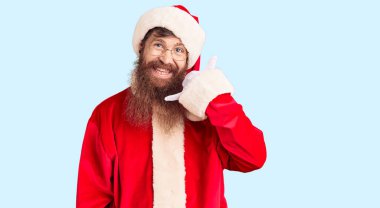 Uzun sakallı, Noel Baba kostümü giyen yakışıklı genç adam telefonla el ve parmaklarıyla telefonla konuşuyor gibi gülümsüyor. iletişim kavramları. 