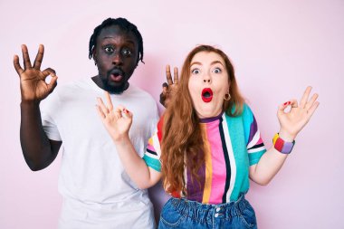 Gündelik kıyafetler giyen ırklar arası bir çift şaşırmış ve şok olmuş görünüyor. Onaylama sembolünü parmaklarıyla yapıyorlar. çılgın ifade 