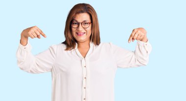 Orta yaşlı, sıradan kıyafetler ve gözlük takan, kendine güvenen, gülümseyen, kendini gururlu ve mutlu gösteren Latin bir kadın.. 