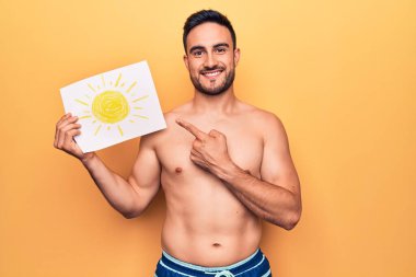 Tatilde sakallı, mayo giyen, elinde güneş ışığı olan, gülümseyen, mutlu, eli ve parmağı olan genç bir adam.