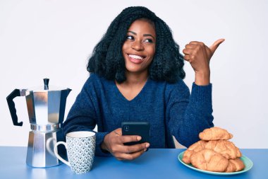 Güzel bir Afrikalı kadın kahvaltısını akıllı telefon kullanarak yapıyor. Başparmağını kaldırıyor, gülümsüyor ve ağzı açık. 