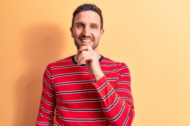 Sıradan çizgili tişört giyen genç yakışıklı adam izole edilmiş sarı arka planda duruyor ve kameraya kollarını kavuşturup çenesine dokunarak gülümsüyor. Olumlu düşünüyorum..