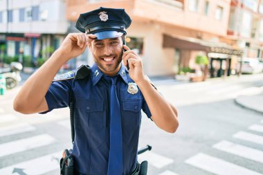 Genç, yakışıklı İspanyol polis memuru polis üniforması giyip mutlu gülümsüyor. Yüzünde tebessümle dikilip şehrin caddesinde akıllı telefondan konuşuyor..