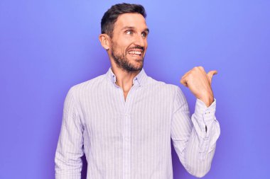 Sıradan çizgili tişört giyen genç yakışıklı, izole edilmiş mor arka planda dikiliyor, baş parmağını yana kaldırıyor ve ağzı açık gülümsüyor.