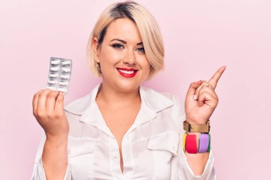 Genç, güzel, sarışın ve cüsseli bir kadın pembe arka planda haplar tutuyor. Gülümsüyor, mutlu bir şekilde işaret ediyor.