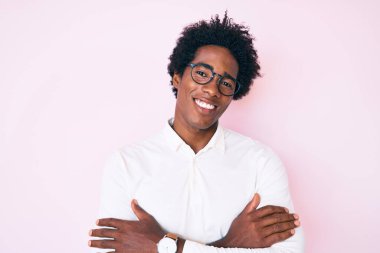 Afro saçlı, iş gömleği giyen ve gözlüklü, kollarını kavuşturup kameraya gülümseyen yakışıklı Afro-Amerikan bir adam. pozitif kişi. 