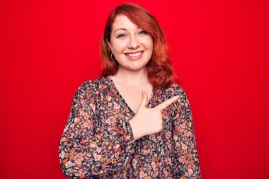 Genç, güzel, kızıl saçlı, çiçekli bir elbise giyen, kırmızı arka planda dikilen, eli ve parmağı yana doğru olan, neşeli gülümseyen bir kadın.