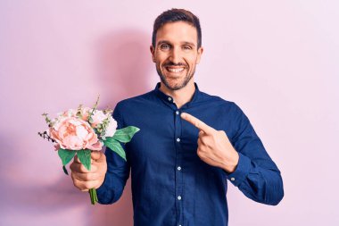 Genç, yakışıklı, romantik bir adam pembe arka planda çiçek buketini tutarken mutlu mesut elini ve parmağını göstererek gülümsüyor.