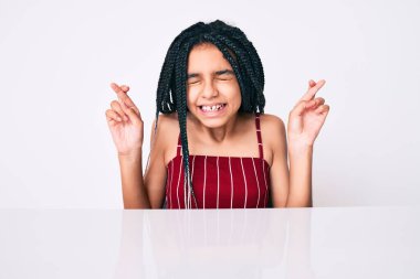 Saçları örgülü genç Afro-Amerikalı kız masanın üzerinde oturmuş umut ve gözlerin kapalı bir şekilde gülümseyerek işaret ediyor. Şans ve batıl inançlar. 