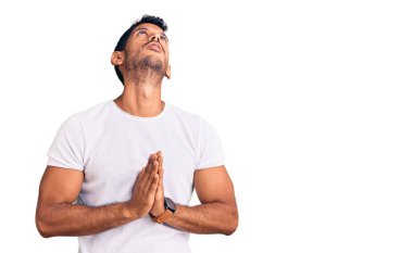 Gündelik kıyafetler giyen genç latin adam elleri bağlı yalvarıyor ve dua ediyor. Yüzünde umut ifadesi var. Çok duygusal ve endişeli. Yalvarıyor. 