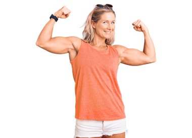 Orta yaşlı, formda, yaz kıyafetleri ve güneş gözlüğü takan, gururla gülümseyen kol kaslarını gösteren sarışın bir kadın. fitness konsepti. 