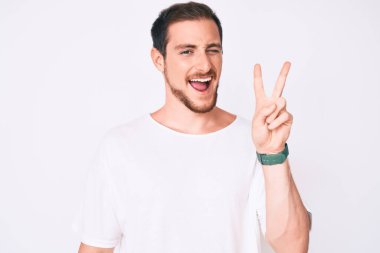 Gündelik beyaz tişört giyen, kameraya gülümseyen ve zafer işareti yapan genç yakışıklı bir adam. İki numara.. 