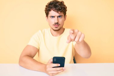 Akıllı telefon kullanarak masanın üzerinde oturan, kamerayı işaret eden beyaz, kıvırcık saçlı genç adam ve sen, kendine güvenen, ciddi görünüyorsun. 