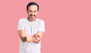 Orta yaşlı yakışıklı, sıradan tişörtler giyen, avuçlarını birbirine değdirip gülümseyen, el hareketi yapan bir adam. Bekleme ve koruma 