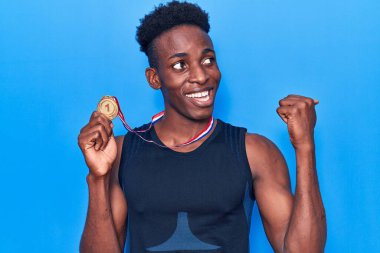 Afro-Amerikalı genç bir adam spor giysisi giymiş elinde madalyayla başparmağını kaldırıp gülümsüyor ve ağzı açık. 