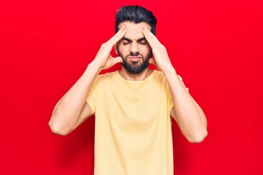 Sakallı, gündelik tişörtlü, baş ağrısı çeken, çaresiz ve stresli genç bir adam. Ağrı ve migren yüzünden. Eller başının üstünde.. 