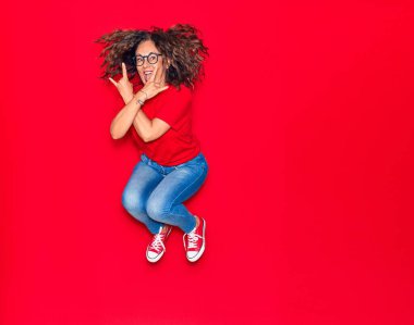 Orta yaşlı, güzel İspanyol bir kadın günlük kıyafetler ve mutlu bir gülümseme takınıyor. İzole edilmiş kırmızı arka planda yüzünde gülümsemeyle zıplıyor