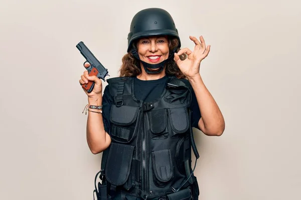 Middle age soldier woman wearing bulletproof uniform and security helmet holding gun doing ok sign with fingers, smiling friendly gesturing excellent symbol