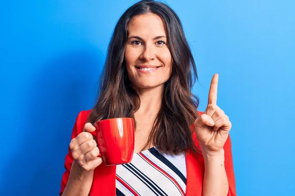 Genç ve güzel esmer iş kadını izole edilmiş mavi arka planda bir kupa kahve içerek bir fikir ya da soruyla mutlu bir yüzü işaret ederek gülümsüyor.