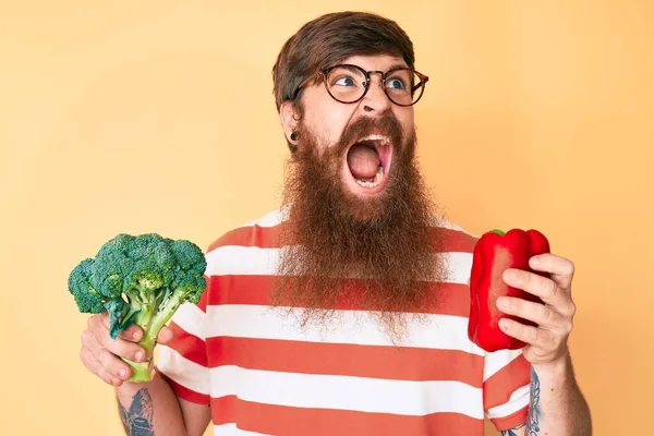 Uzun sakallı, elinde brokoli ve kırmızı biber olan yakışıklı, genç ve kızıl saçlı bir adam sinirli ve öfkeliydi. Öfkeyle bağırıyordu. öfke ve agresif konsept. 