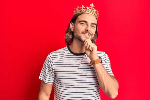 Young handsome man wearing prince crown smiling looking confident at the camera with crossed arms and hand on chin. thinking positive.