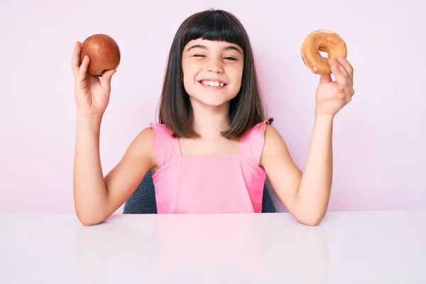 Маленькая Девочка Бумом Держа Красное Яблоко Пончик Сидя Столе Подмигивая — стоковое фото