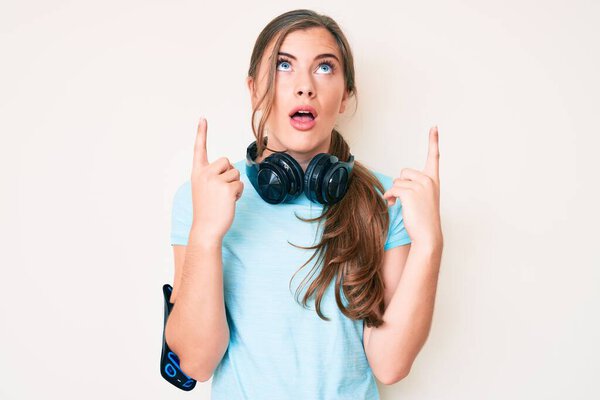 Красивая молодая белая женщина в спортивной одежде и наушниках удивлена и удивлена, глядя вверх и указывая пальцами и поднимая руки.. 