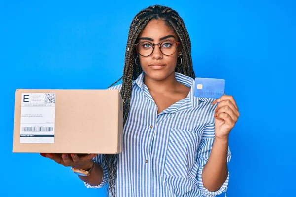 Saçları örgülü, kredi kartı olan, yüzünde ciddi bir ifadeyle rahatlamış genç bir Afrikalı Amerikalı kadın. Basit ve doğal bir şekilde kameraya bakıyorsun.. 