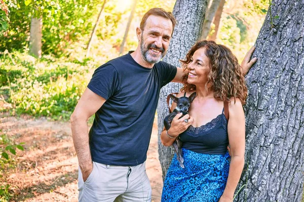 Orta yaşlı çift gülümsüyor. Chihuahua köpeği kucağında. Ormandaki kameraya bakıyor.
