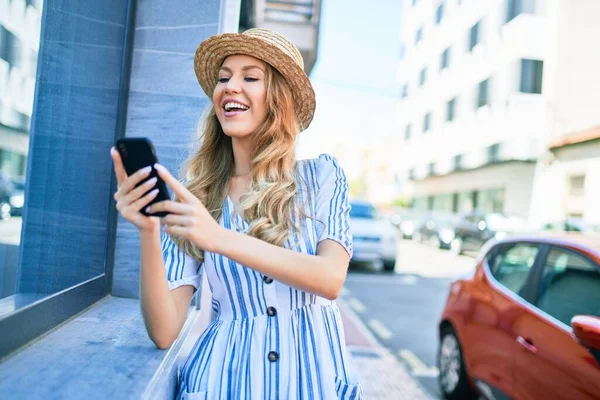 年轻美丽的金发女子戴着夏帽在度假时快乐地笑着 在城市街道上用智能手机笑着靠在墙上 — 图库照片