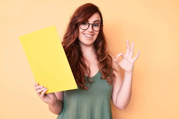 Young beautiful woman wearing glasses holding book doing ok sign with fingers, smiling friendly gesturing excellent symbol