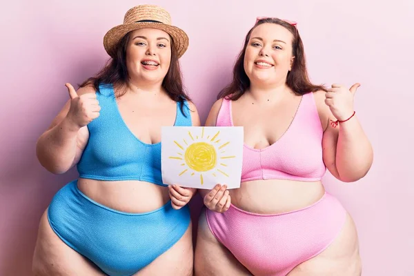 Young plus size twins wearing bikini holding sun draw pointing thumb up to the side smiling happy with open mouth