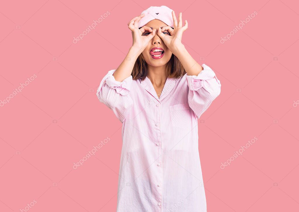 Young beautiful chinese girl wearing sleep mask and pajama doing ok gesture like binoculars sticking tongue out, eyes looking through fingers. crazy expression. 