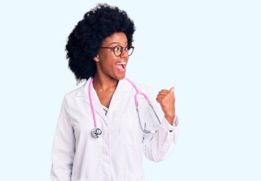 Doktor önlüğü ve steteskop giymiş, gülümseyen, gülümseyen ve baş parmağıyla yüzünü gösteren Afro-Amerikan genç bir kadın.. 