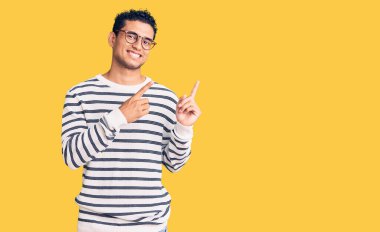 Latin asıllı yakışıklı genç adam gündelik kıyafetler ve gözlük takıyor gülümsüyor ve kameraya bakıyor iki eliyle ve parmaklarıyla kenarı işaret ediyor.. 