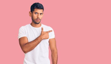 Gündelik kıyafetler giyen genç latin adam, el parmağı ile yan tarafı işaret ederek ciddi ve sakin bir yüz ifadesi sergiliyor. 