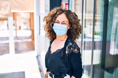 Orta yaşlı güzel esmer kadın tıbbi maske takıyor teras restoranında duruyor.