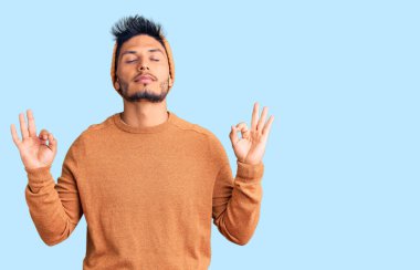 Yakışıklı Latin Amerikalı süveter ve yün şapka giyen, gözleri kapalı gülümseyen, parmaklarıyla meditasyon yapan genç adam. yoga konsepti. 